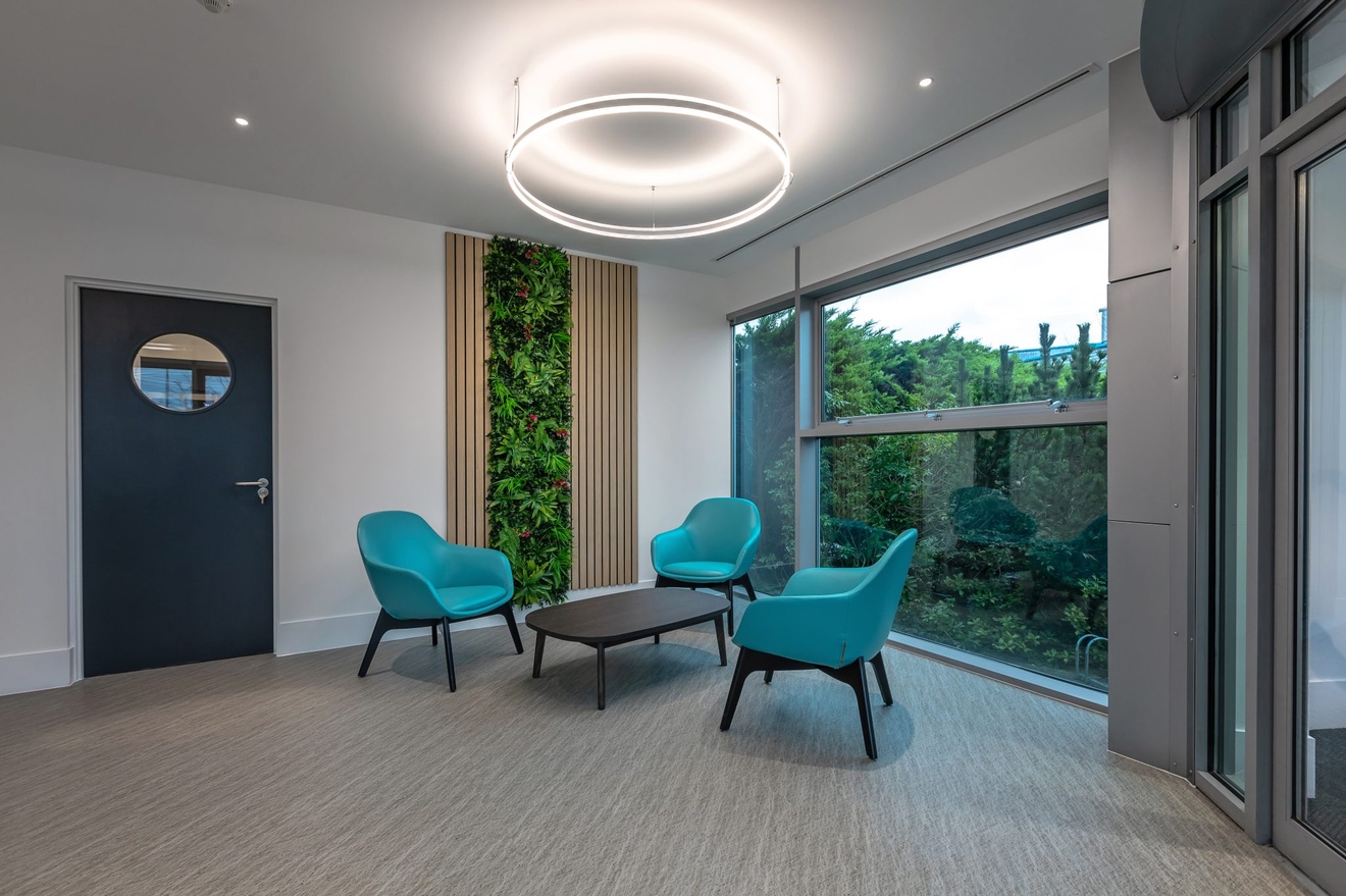 small waiting area with black coffee table and blue chairs with large windows leading to green trees